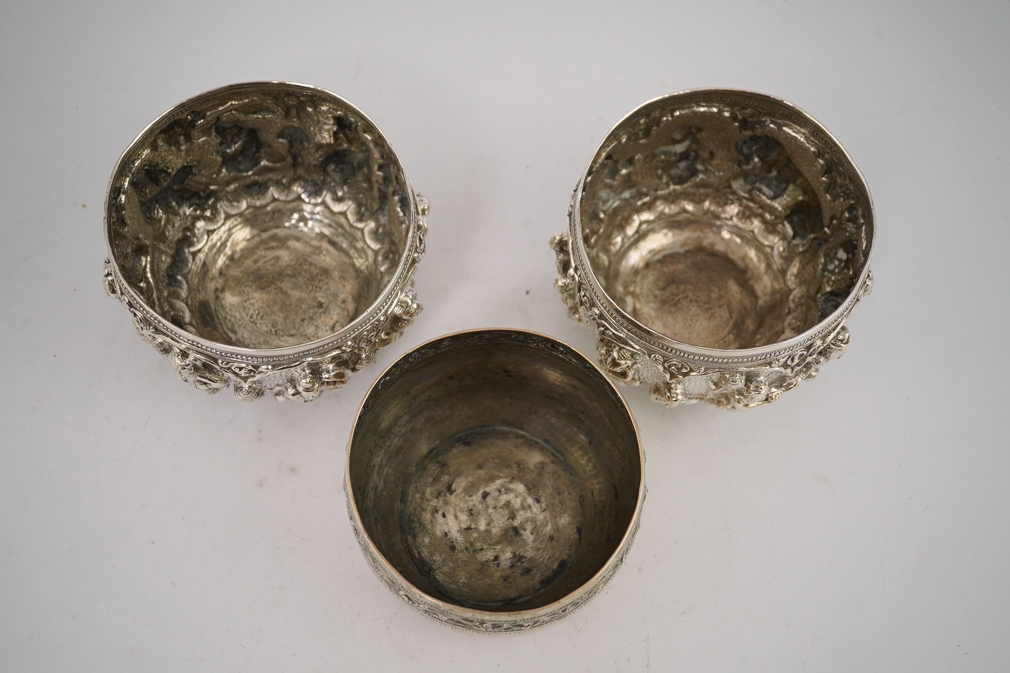 A pair of Burmese white metal bowls, embossed with figures, 12cm diameter, 220 and 224g and a plainer bowl, 110g. Condition – fair